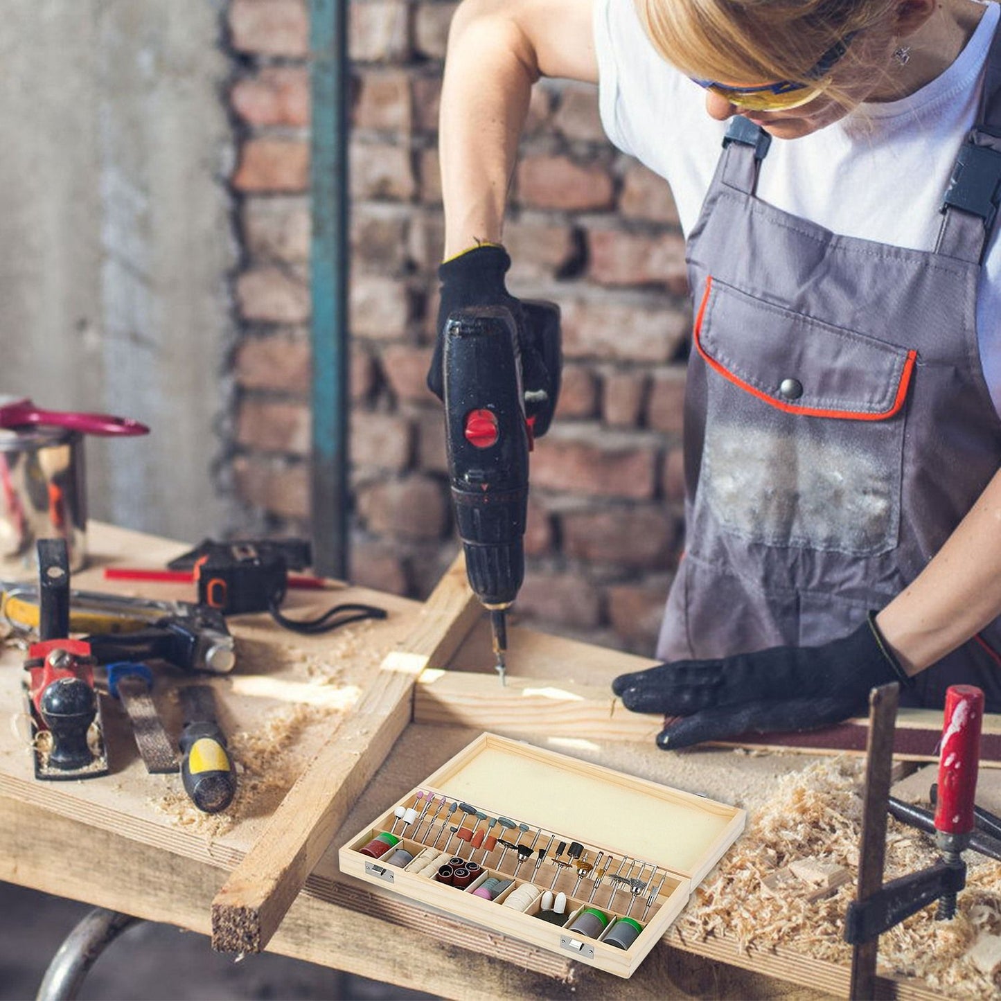 Woman working on a woodworking project with a power drill, surrounded by tools and a wooden box of colorful thread spools on a workbench.