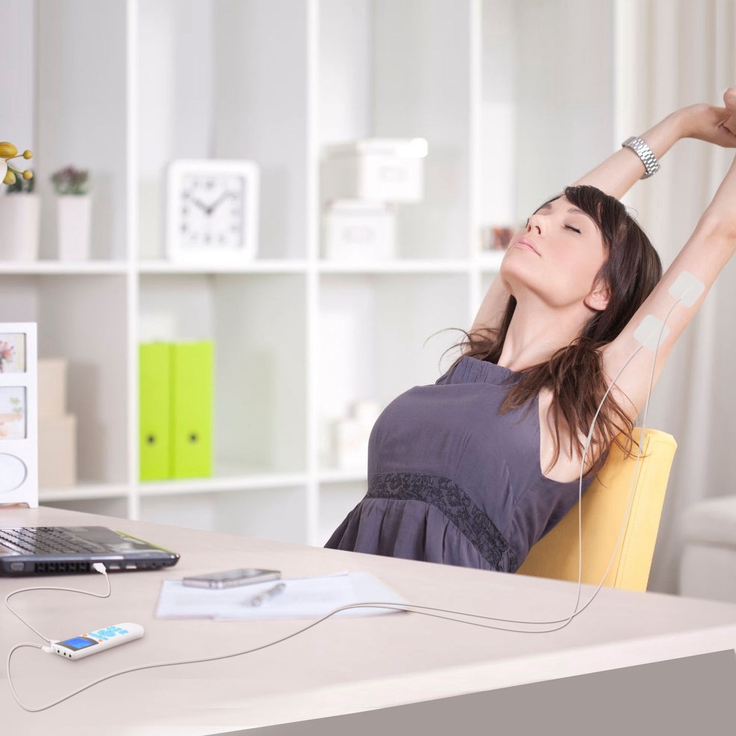 Woman using a portable electronic muscle stimulator on her arm while seated at a desk, promoting relaxation and pain relief in a stylish home office environment.