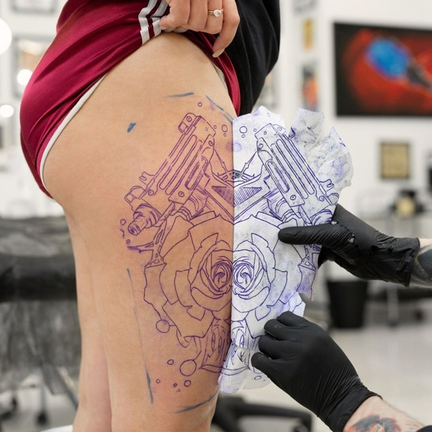 Tattoo artist transferring a detailed design of guns and roses onto a client's thigh, showcasing the artistic process in a tattoo studio setting.