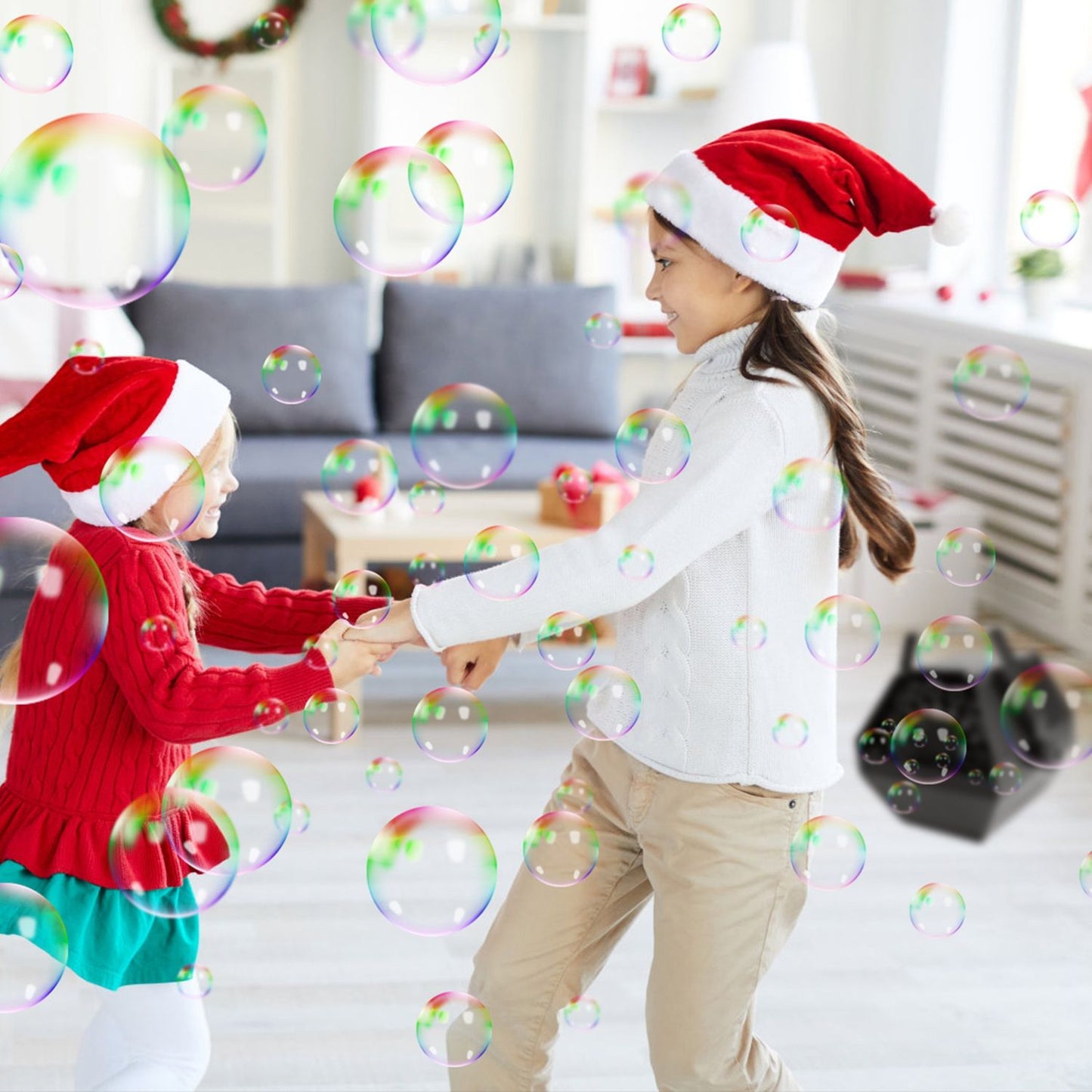 Two children wearing festive Santa hats playfully dance together amidst floating bubbles in a bright, cozy living room decorated for the holidays.
