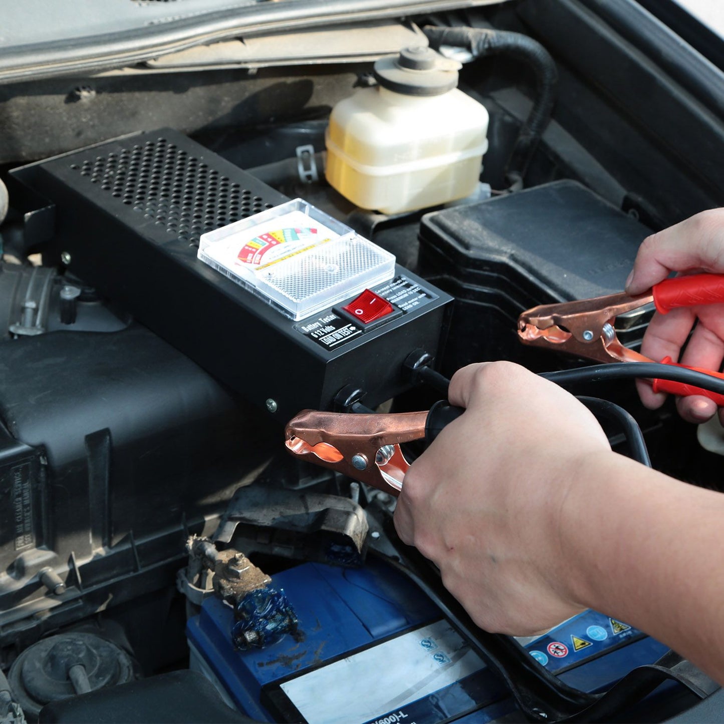 A person uses jumper cables to connect a portable car battery jump starter to a vehicle's battery under the hood, showcasing the tool's functionality in a car maintenance setting.