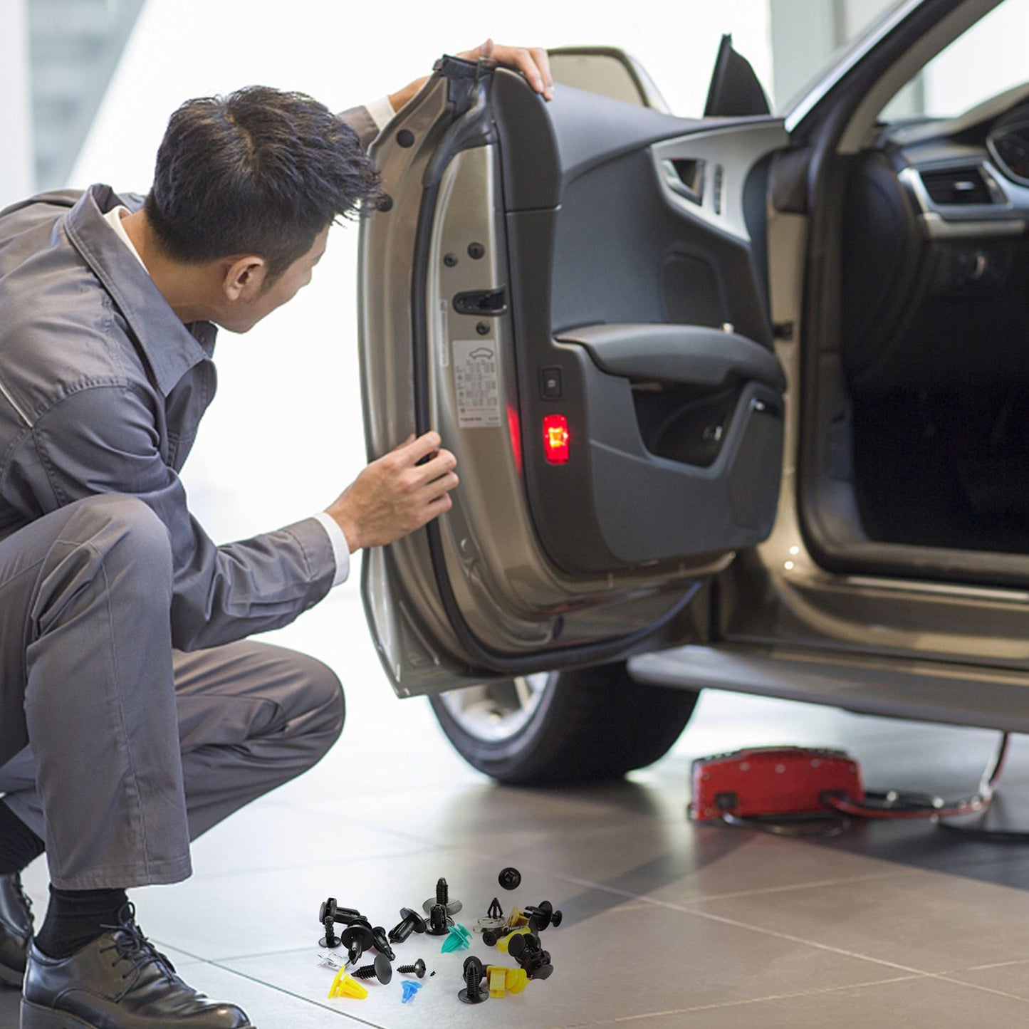 Automotive technician inspecting a car door, surrounded by various car fasteners and clips on the floor, in a well-lit workshop setting.