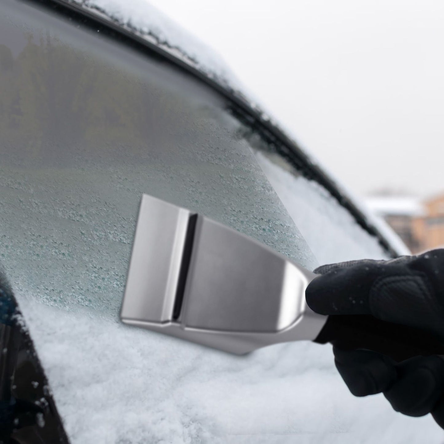 Person using a stainless steel ice scraper to remove frost and snow from a car windshield on a chilly winter day.