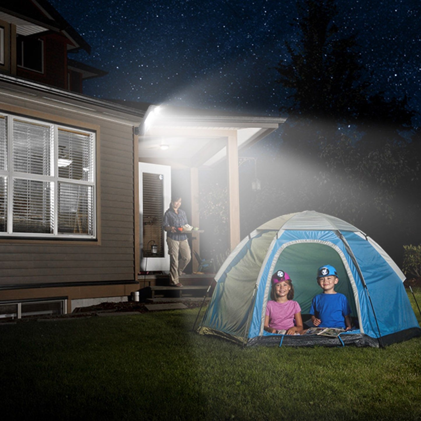 Family camping in the backyard at night, featuring a colorful tent set on green grass with two children inside, while a parent stands on the porch with a tray, under a starry sky.