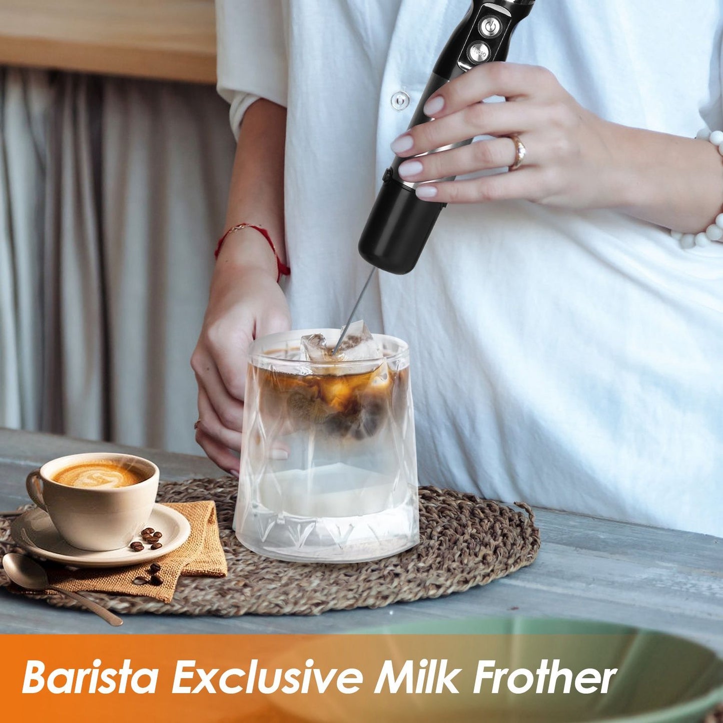 A person using a black Barista Exclusive Milk Frother to froth milk in a transparent glass with iced coffee, and a cup of espresso sits on a rustic table.