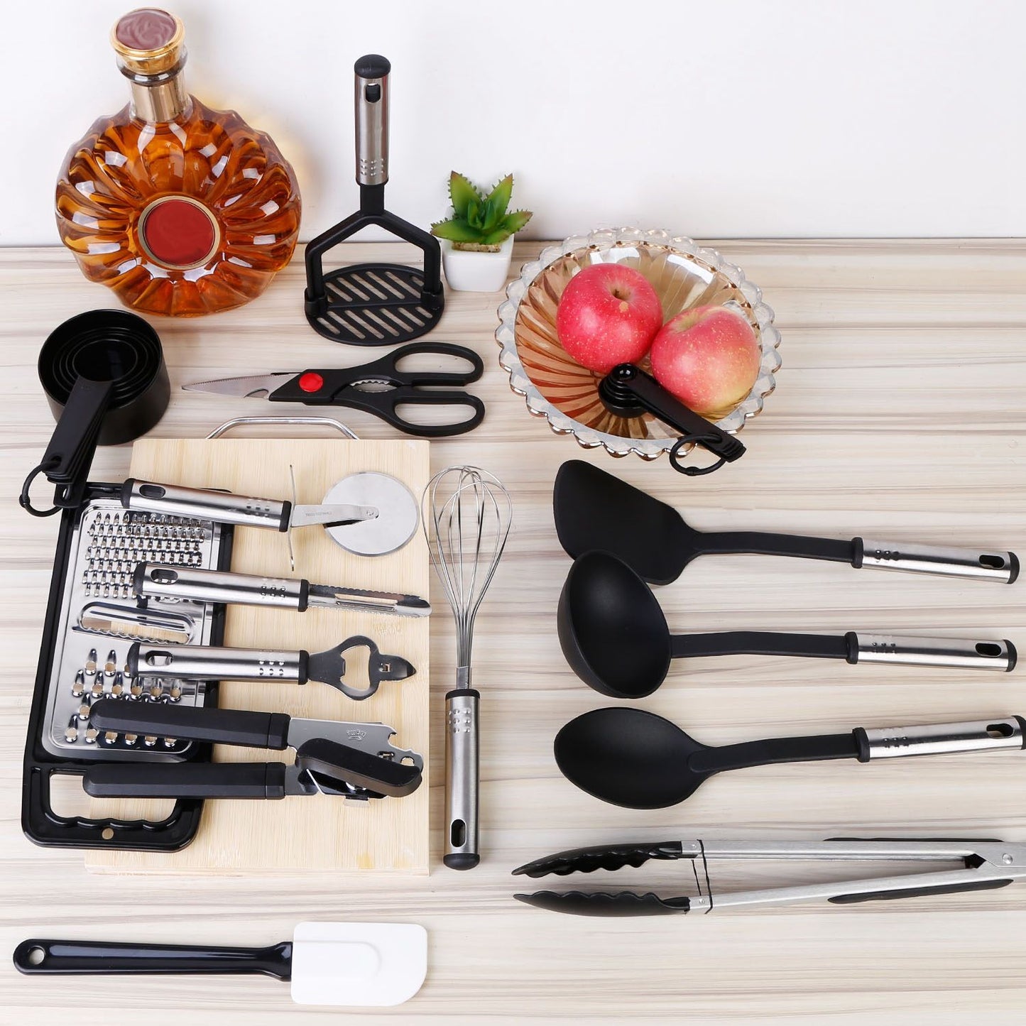 Kitchen utensils set featuring various cooking tools such as measuring cups, spatula, tongs, and a whisk, arranged on a wooden cutting board alongside apples, a decorative bowl, and a bottle of whiskey, all on a light wooden surface.
