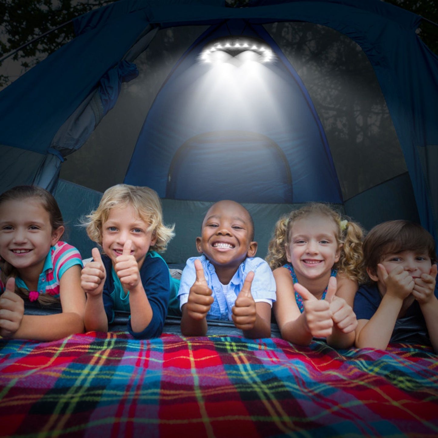 Group of happy children giving thumbs up inside a brightly lit tent, with a colorful plaid blanket underneath, perfect for family camping adventures.
