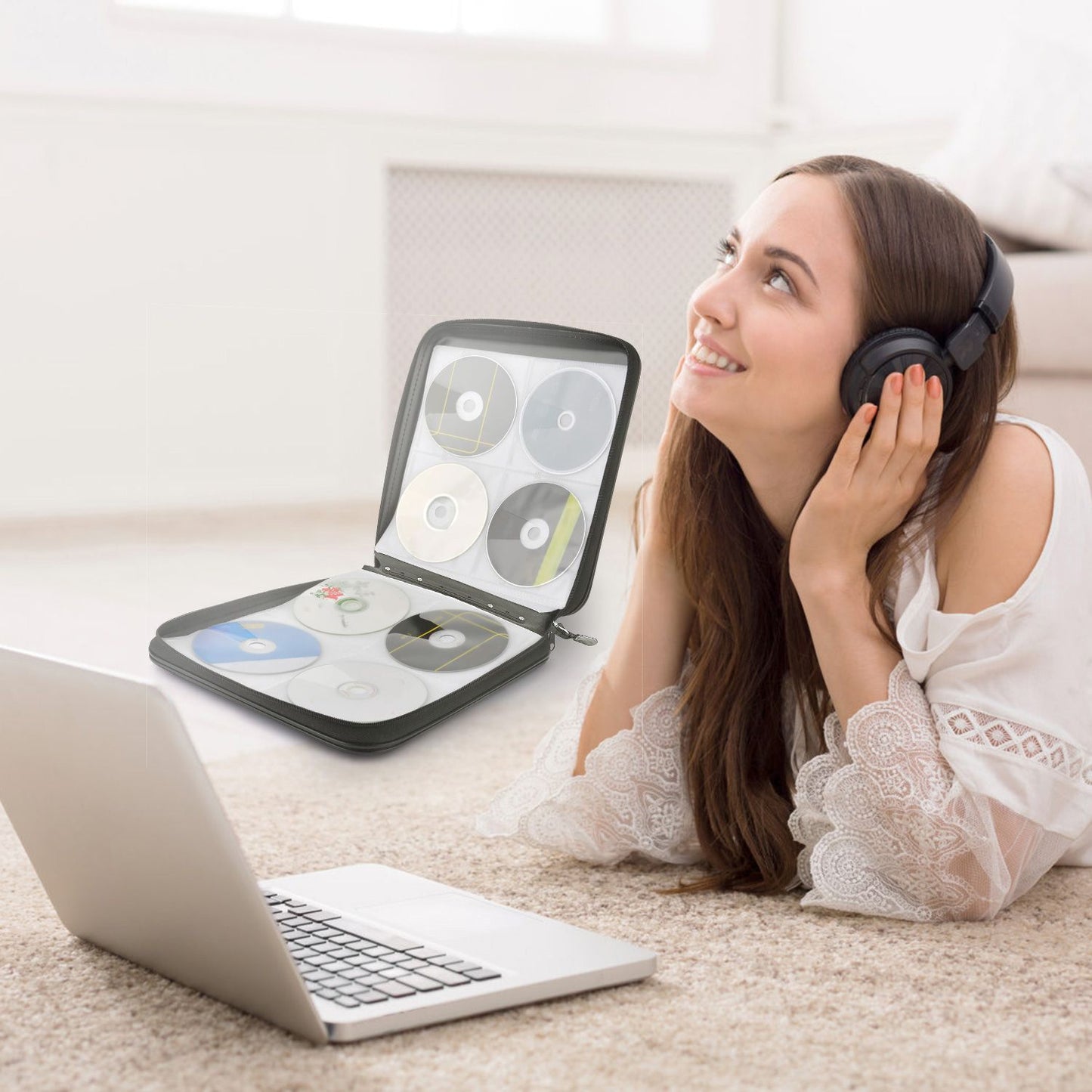 CD case organizer opened with CDs displayed next to a woman listening to music on headphones, relaxing on a carpet with a laptop.