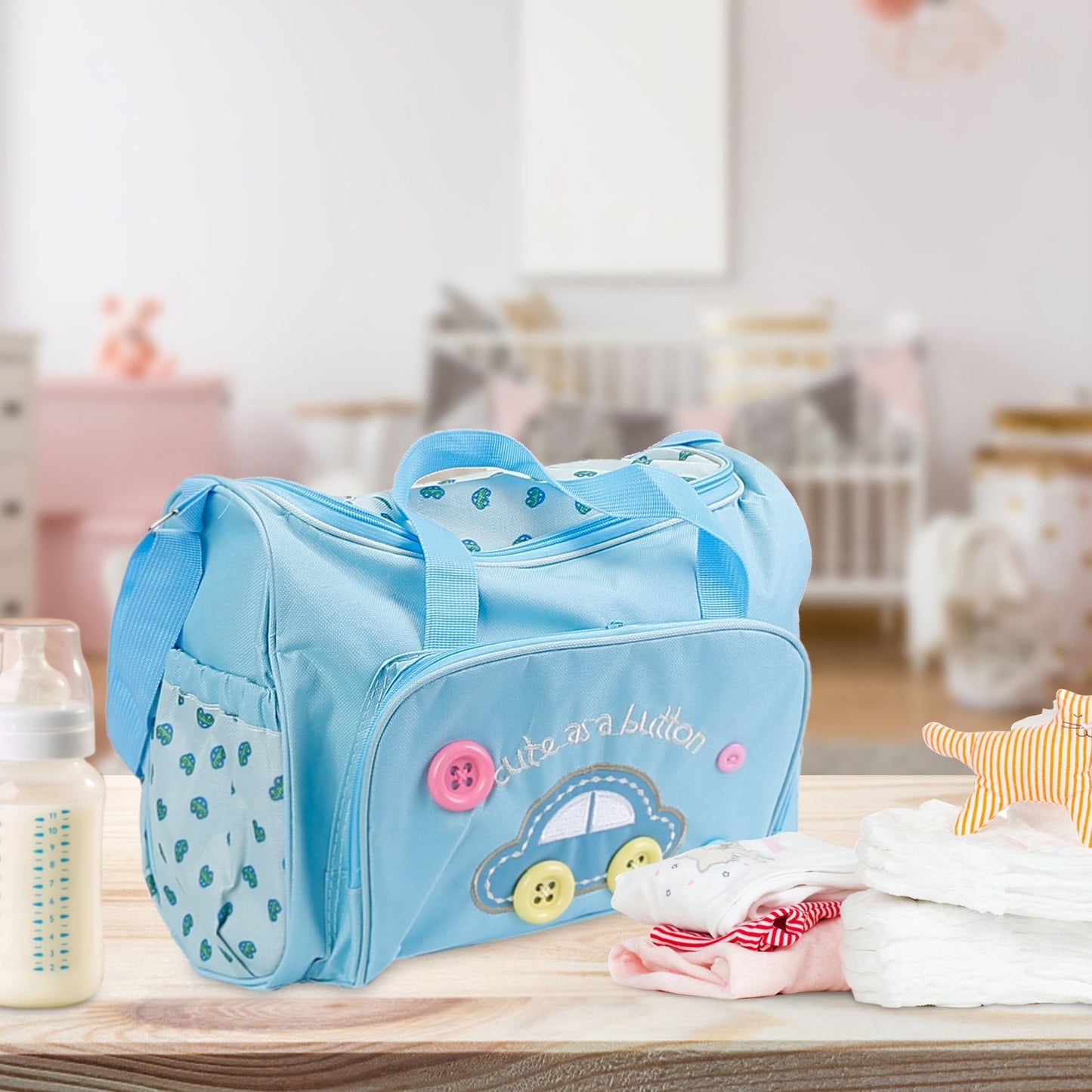 Light blue baby bag with playful car design, buttons, and the phrase "cute as a button," accompanied by a baby bottle and neatly folded baby clothes on a wooden surface in a cozy nursery setting.