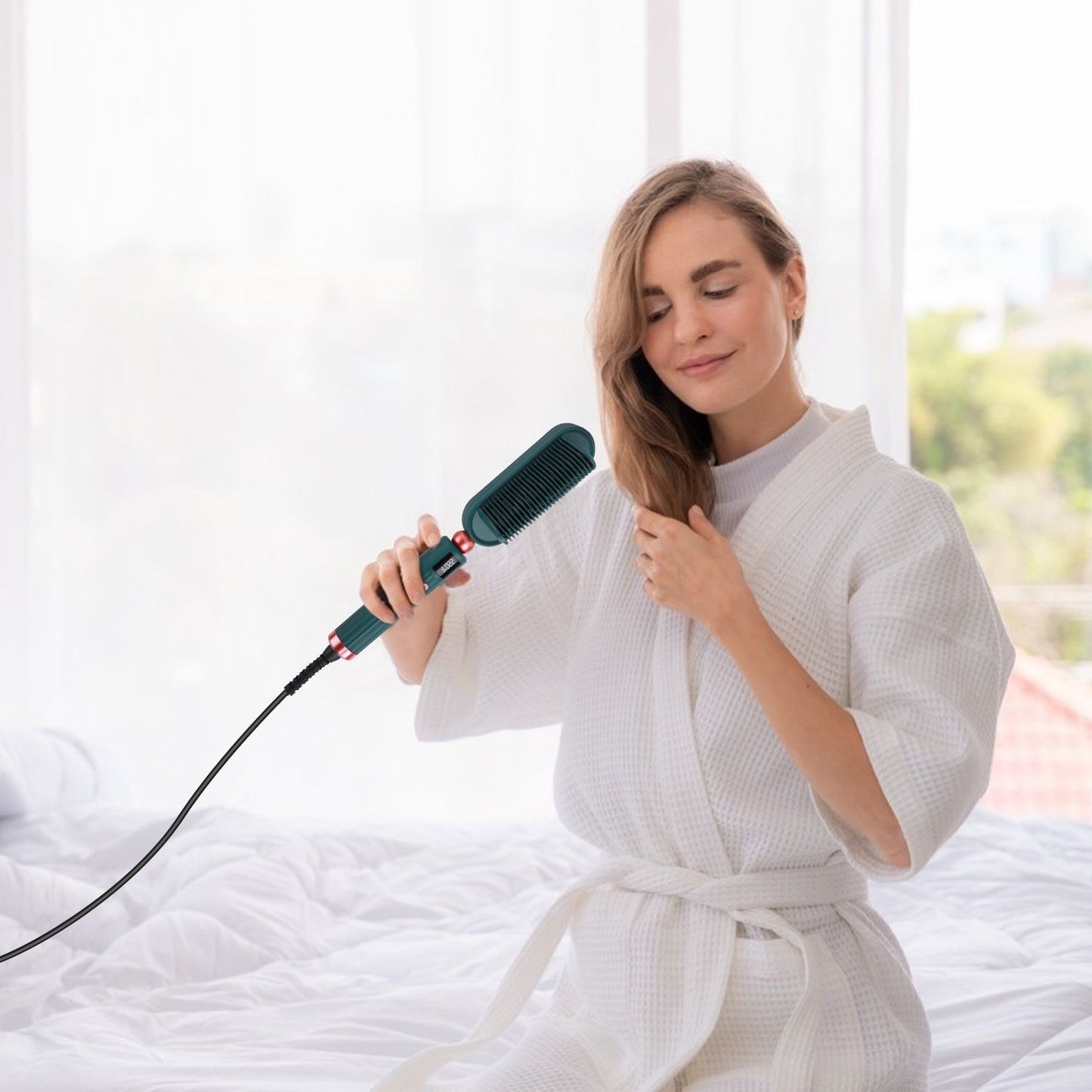 Model using a teal heated hairbrush while wearing a white bathrobe, sitting on a bed with soft bedding and natural light coming through a window.