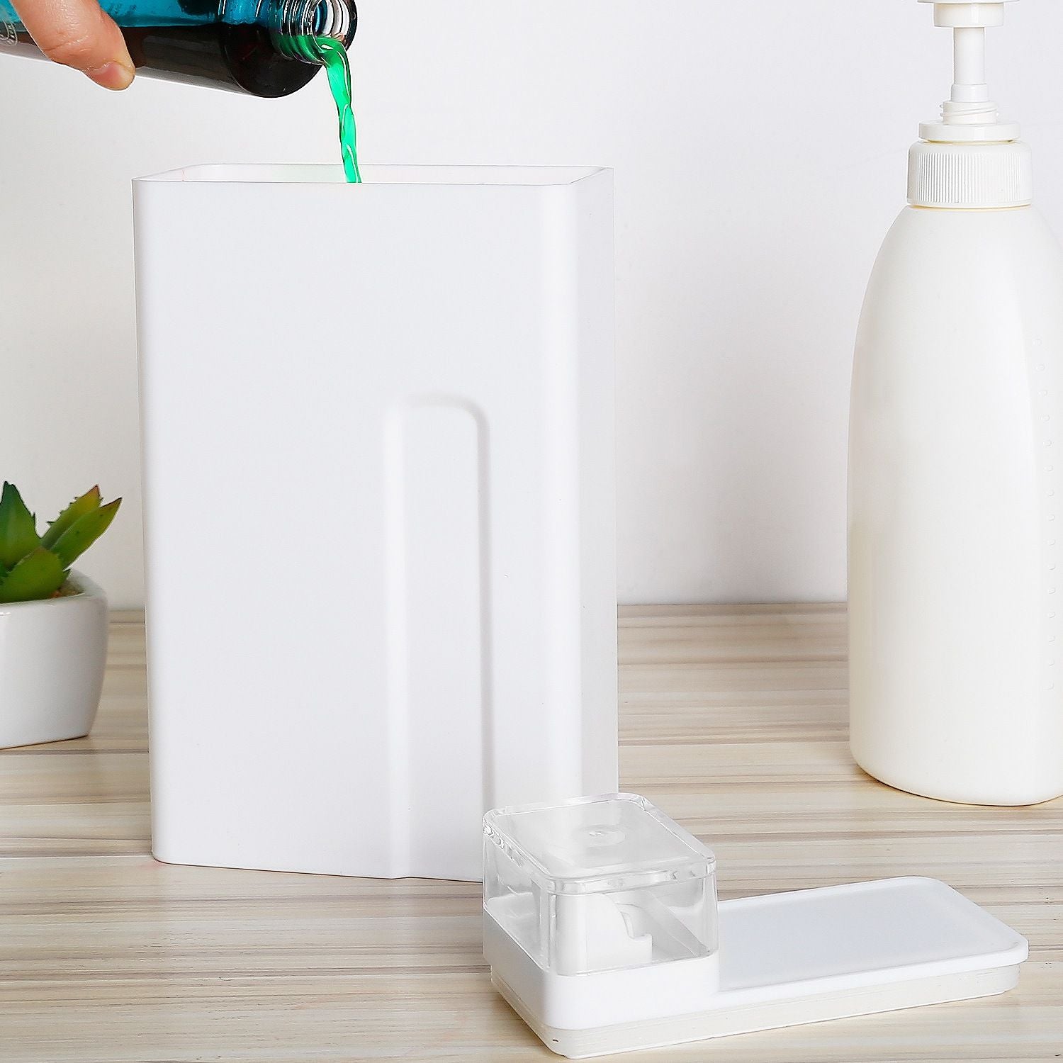 White rectangular container with a green liquid being poured in, alongside a clear soap dispenser and a small white dish, on a wooden surface with a small plant in a pot.
