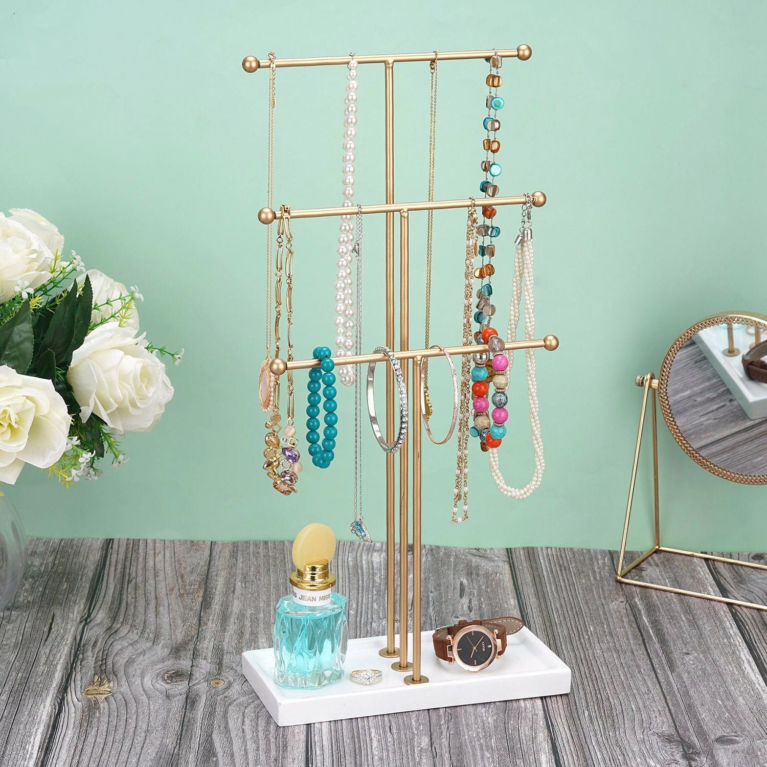Elegant gold jewelry organizer stand displaying various necklaces and bracelets, alongside a perfume bottle and wristwatch, with soft white roses in the background.