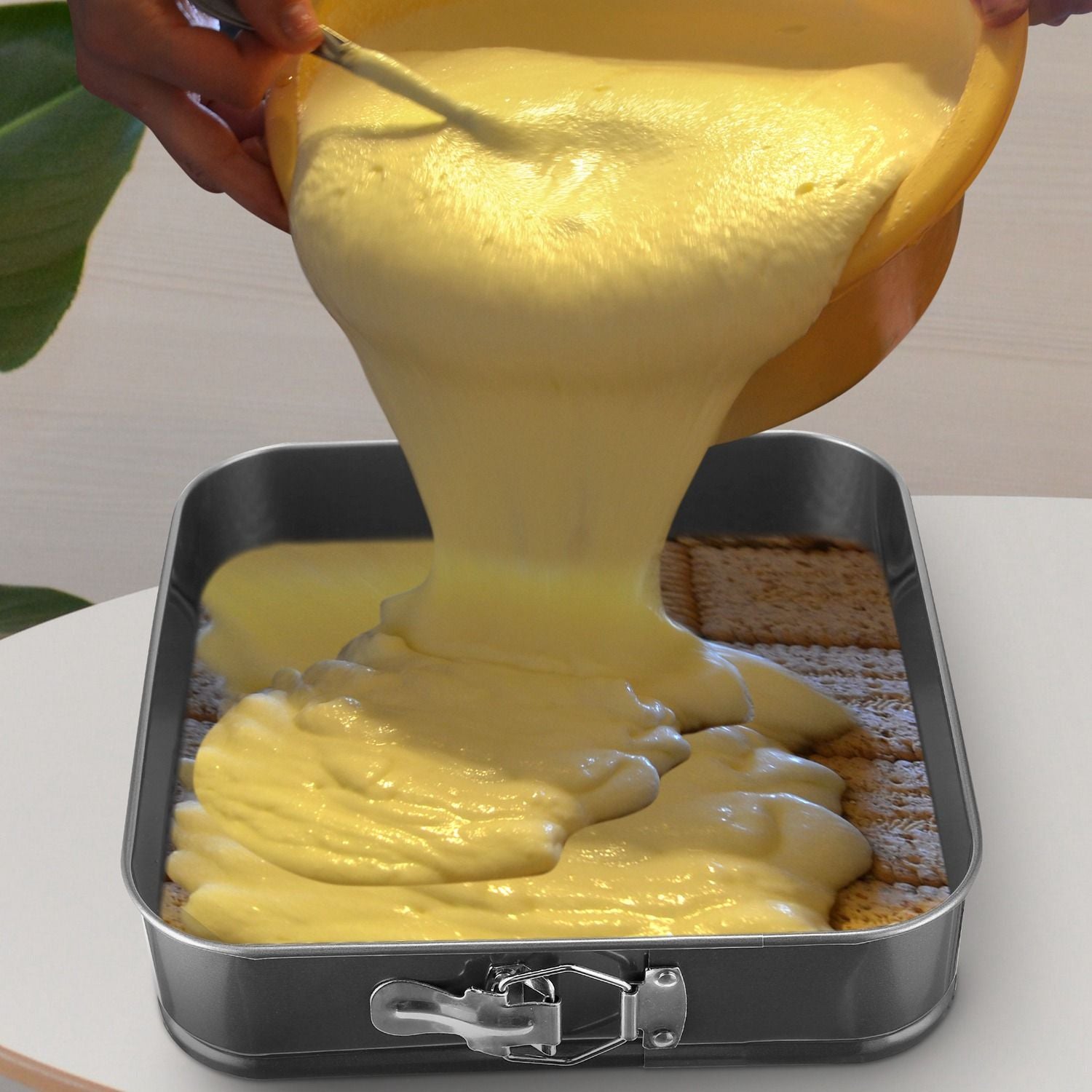 Person pouring creamy yellow batter into a rectangular baking dish lined with graham crackers, with a focus on the smooth texture of the mixture and the baking pan's stainless steel edge.
