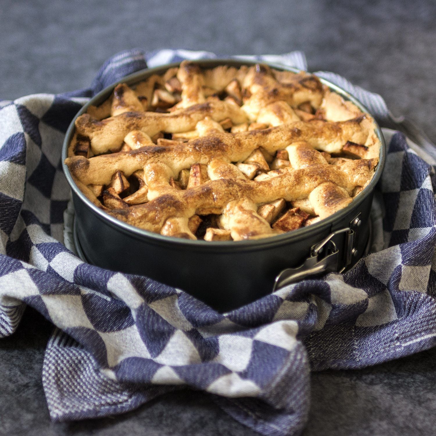 Freshly baked apple pie in a round springform pan, with a golden brown lattice crust, resting on a blue and white checkered kitchen towel.