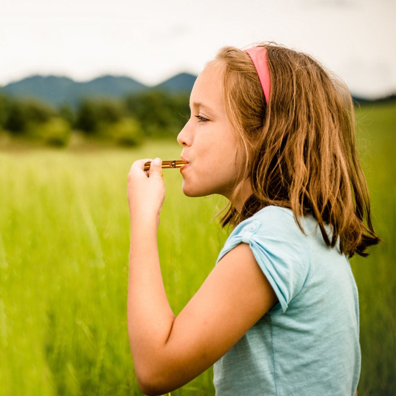 Girl holding a key in a field of green grass with mountains in the background, wearing a light blue shirt and a pink headband.