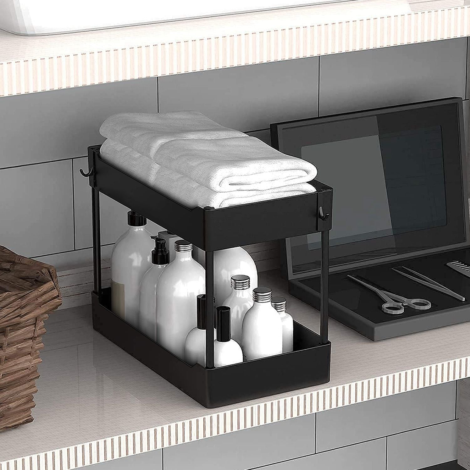 Black metal bathroom storage organizer with neatly folded towels on top and various white bottles below, placed on a modern vanity countertop with a basket and grooming tools.