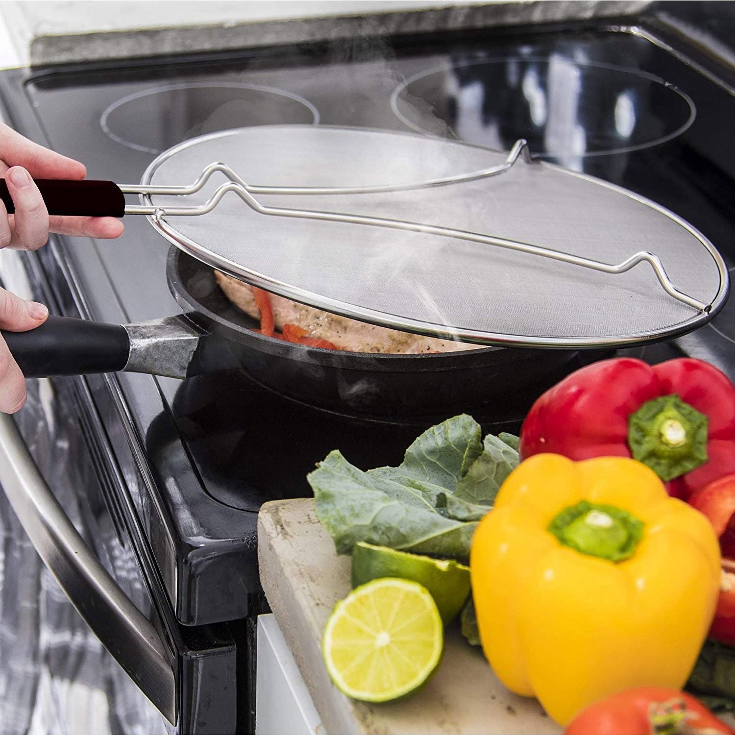 Stainless steel splatter guard being used on a frying pan with steaming food, surrounded by fresh vegetables including bell peppers and lime.