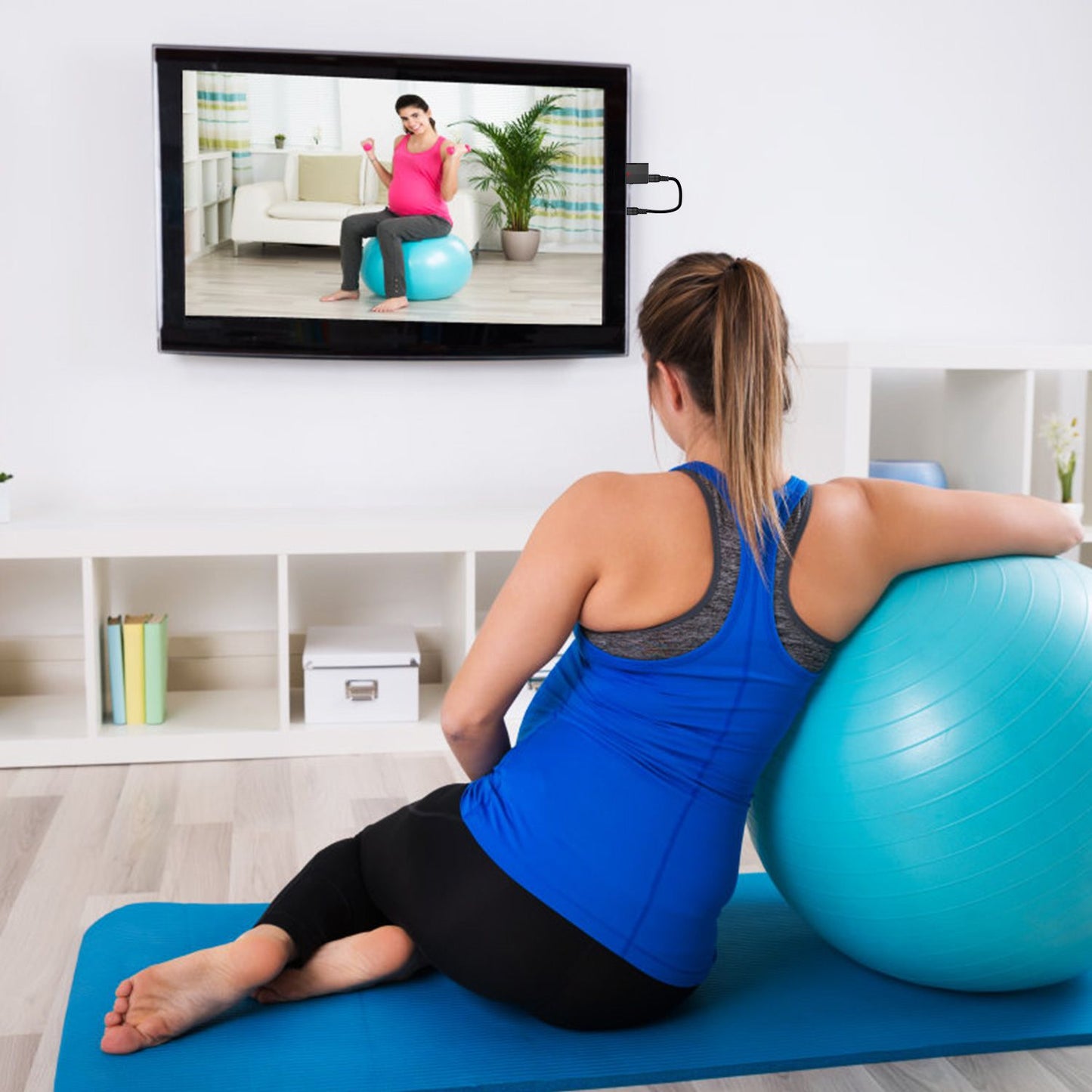 Pregnant woman exercising with a stability ball at home, following a workout video on television, promoting fitness and wellness during pregnancy.