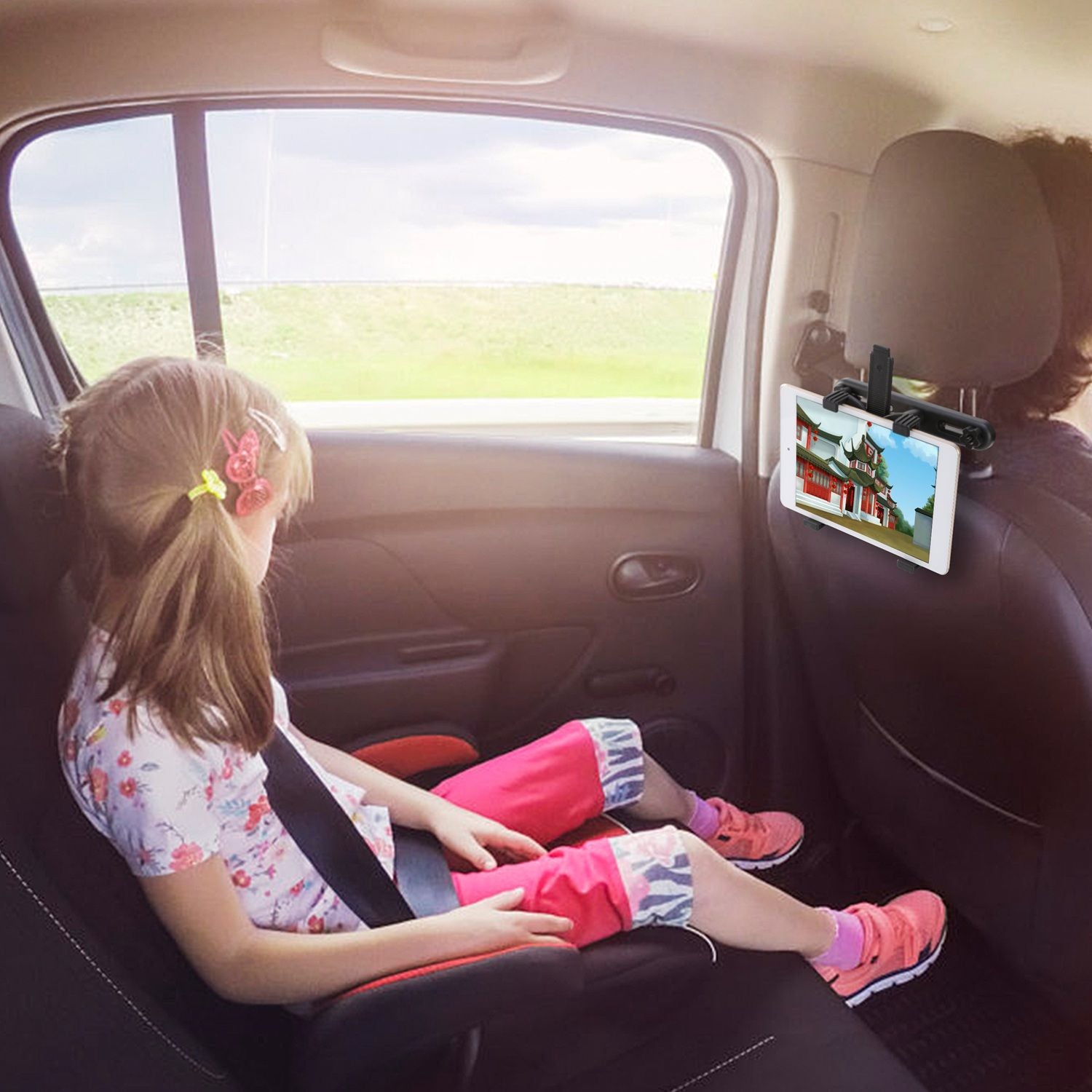 A young girl with hair clips sits in the back seat of a car, wearing a floral shirt and colorful pants, while watching a tablet mounted on the headrest, providing entertainment during the ride.