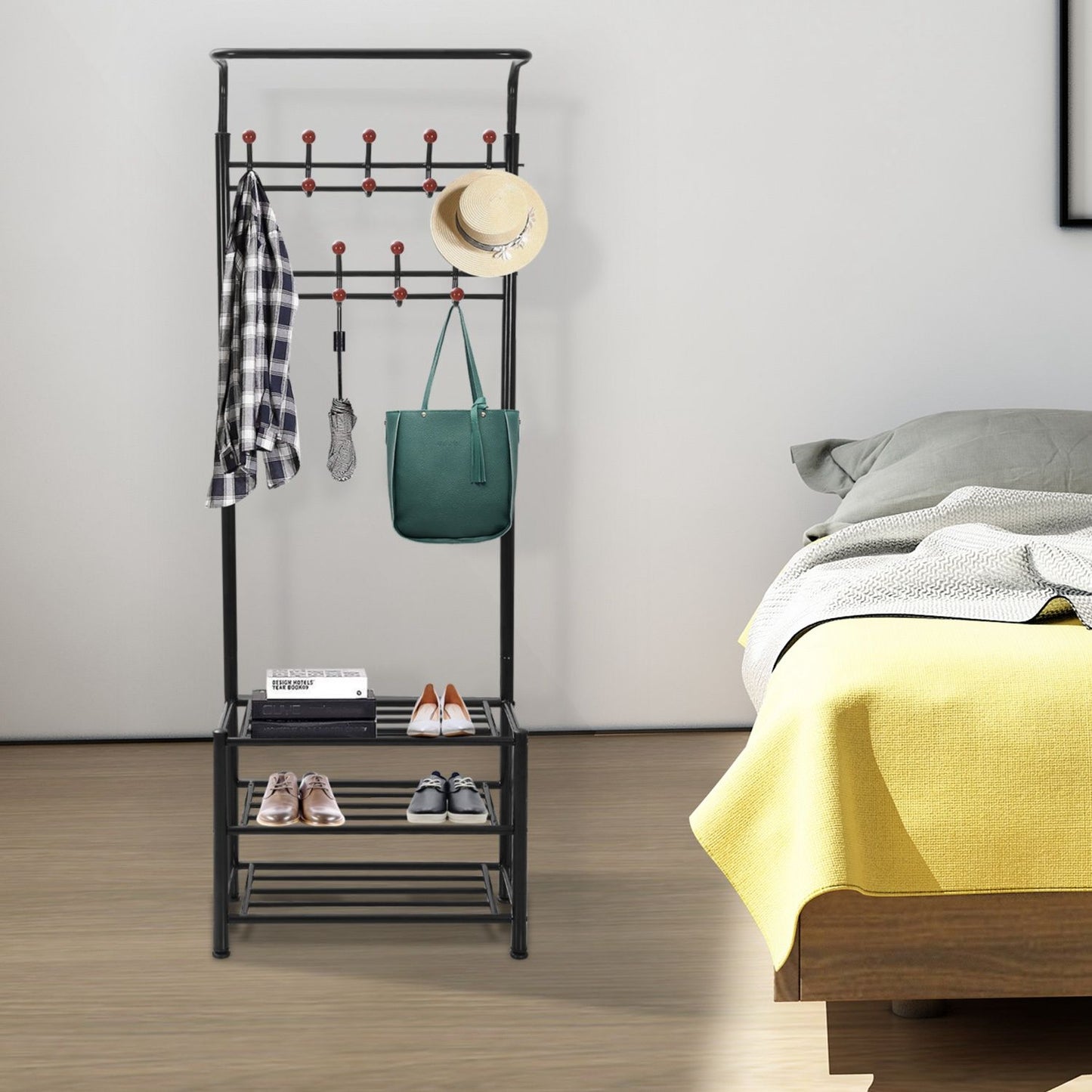 Black metal coat rack with multiple hooks, a green handbag, a hat, and neatly arranged shoes on a shelf, positioned beside a neatly made bed with a yellow throw blanket.