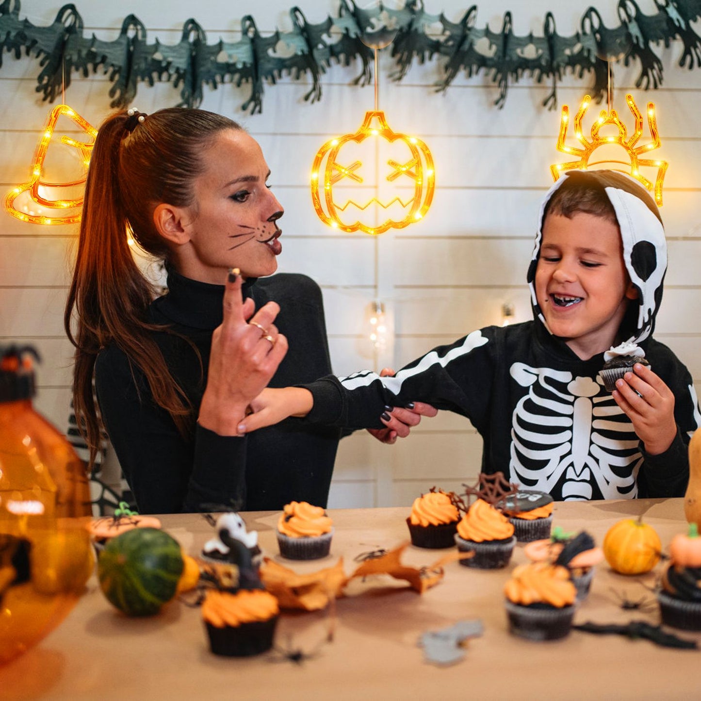 A woman and a child dressed in a skeleton hoodie celebrate Halloween at a festive table filled with spooky-themed cupcakes, pumpkins, and decorations, with glowing Halloween lights in the background.