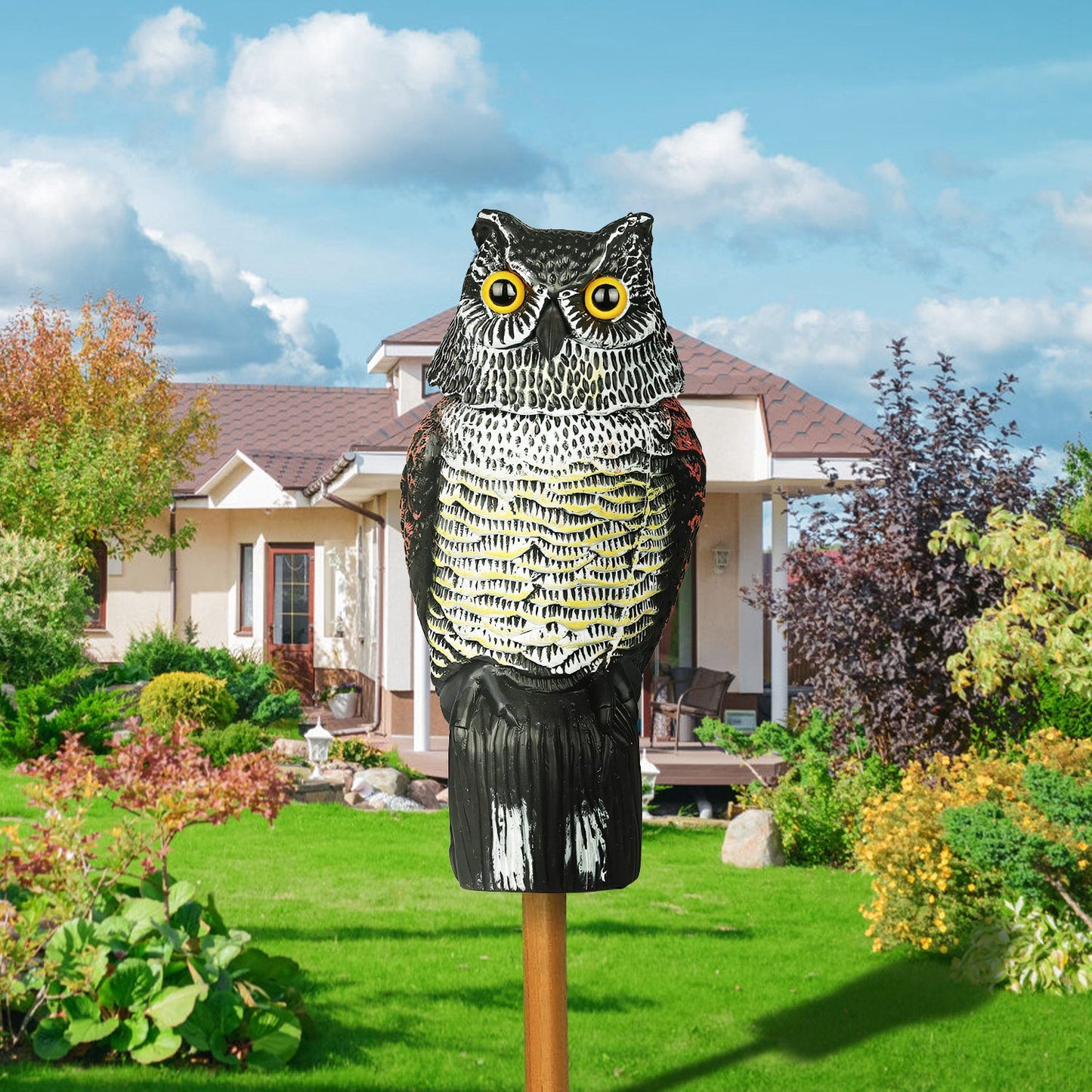 Realistic owl garden decoy with yellow eyes, designed to scare away birds and pests, set in a vibrant green landscape with a house in the background.