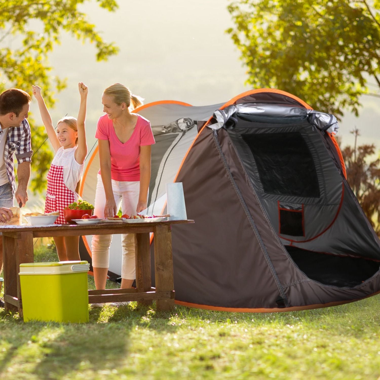 Family enjoying a summer camping trip, preparing food at a wooden table with a green cooler, in front of a spacious, gray camping tent set in a sunny outdoor setting surrounded by trees.