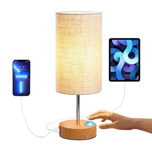 Modern bedside lamp with USB charging ports, featuring a soft fabric shade, wooden base, and touch sensor, with a smartphone and tablet connected for charging.