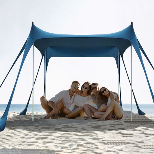 Group of friends relaxing under a stylish blue beach tent on a sandy shore with ocean in the background.
