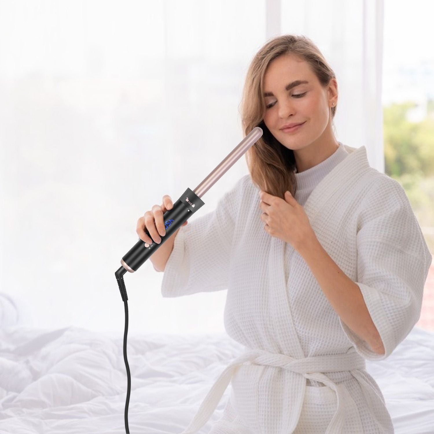 Woman in a white bathrobe using a hair curling wand to style her long hair at home, showcasing the sleek design and functionality of the curling iron.