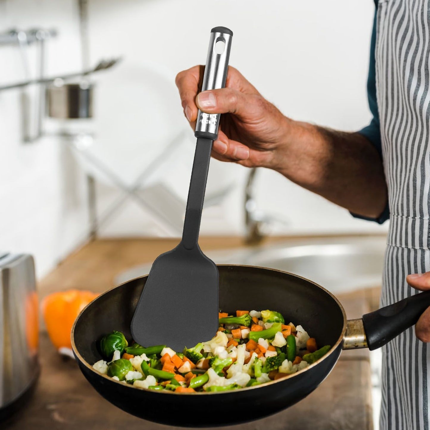 A person using a silicone spatula to stir stir-fried vegetables in a black pan on a wooden kitchen countertop.
